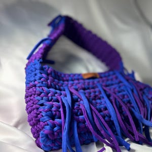 May include: A handmade crochet bag in shades of blue and purple. The bag features a woven design with a fringe detail. The strap is a solid purple, and the fringe is a mix of blue and purple. The bag is set against a white background.