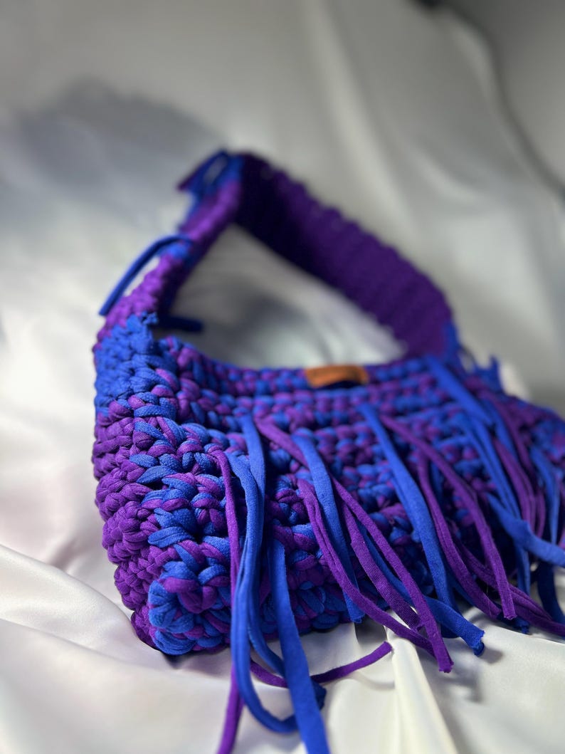 May include: A handmade crochet bag in shades of blue and purple. The bag features a woven design with a fringe detail. The strap is a solid purple, and the fringe is a mix of blue and purple. The bag is set against a white background.