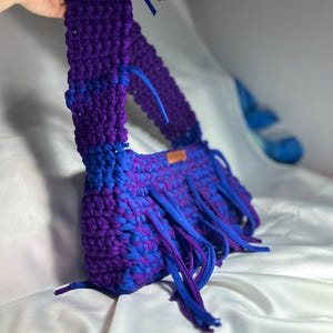 May include: A handmade, crochet bag in shades of blue and purple. The bag features a long strap and fringe detailing along the bottom edge. The bag is made of thick, soft yarn, and the colours are blended throughout the design.