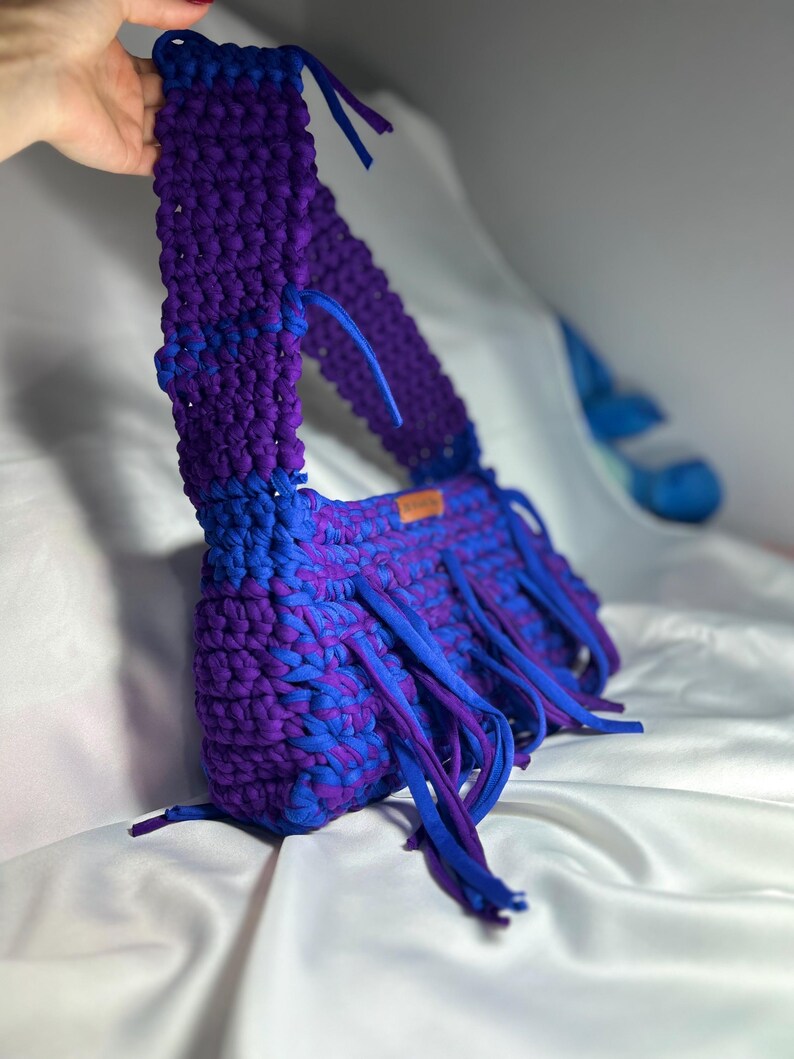 May include: A handmade, crochet bag in shades of blue and purple. The bag features a long strap and fringe detailing along the bottom edge. The bag is made of thick, soft yarn, and the colours are blended throughout the design.
