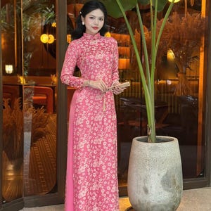 May include: A pink long-sleeved dress with a white floral pattern. The dress has a mandarin collar and a long skirt. The model is standing in front of a glass door and a potted plant.