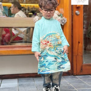 May include: A young boy wearing a light blue traditional Vietnamese tunic with a dragon and wave pattern. He is wearing glasses and blue jeans with distressed details. The tunic has a mandarin collar and long sleeves. The boy is standing in front of a wooden door with a sign that says "Ao Dai UK".