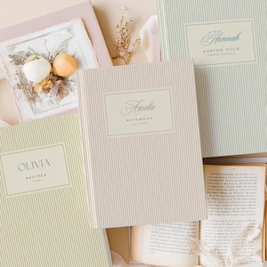 Pode incluir: Uma coleção de cadernos e diários personalizados com capas listradas em várias cores, incluindo verde-claro, rosa e creme. Cada caderno apresenta um nome ou título personalizado em escrita elegante dentro de uma moldura retangular. Um caderno está rotulado como "Olivia Recipes".