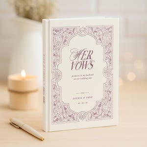 May include: A cream-colored wedding vow book with the words "Her Vows" in a decorative font. The cover features an ornate, purple border and the names "Sophia & Dave" with a date. A pen and candle are in the background.