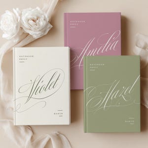 Op de afbeelding: Drie notitieboeken in roze, crème en groen. Elk notitieboek bevat een naam in elegant schrift: "Amelia", "Violet" en "Hazel". De notitieboeken zijn gelabeld met "Notebook, property of" en "March 2027".