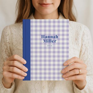 May include: A hardcover notebook with a blue spine and a light purple and white gingham pattern. The cover features the name "Hannah Miller" and the year "2026" in a dark blue font. The notebook is held in front of a person.