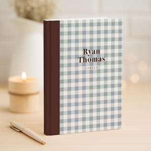 May include: A hardcover notebook with a brown spine and a light blue and white gingham pattern. The cover features the name "Ryan Thomas" and the word "Lyrics" in a dark brown font. A white pen rests on the wooden surface.