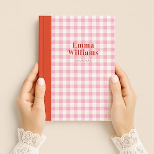 May include: A journal with a pink and white gingham pattern cover and a red spine. The text "Emma Williams JOURNAL" is printed on the cover in a serif font. The journal is held by two hands.