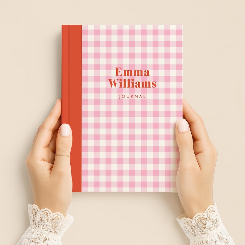 May include: A journal with a pink and white gingham pattern cover and a red spine. The text "Emma Williams JOURNAL" is printed on the cover in a serif font. The journal is held by two hands.