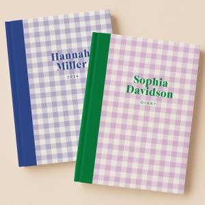 May include: Two hardcover journals with a gingham pattern in lavender and white. One journal has a blue spine and the name "Hannah Miller" and the year "2026" printed on the cover. The other journal has a green spine and the name "Sophia Davidson" and the word "DIARY" printed on the cover.