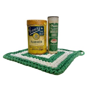 May include: A yellow cylindrical Cook's The Best Flaxseed tin and a tall, thin Phenix Vegetable Bouillon Cubes container resting on a square, green and white crocheted potholder with a small loop on one corner for hanging purposes.