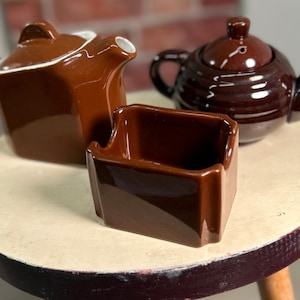 May include: Three brown ceramic teaware pieces sit on a small, round table. The set includes two teapots, one square and one round, and a small rectangular container. The teapots have lids and spouts, and the square teapot has a white rim.