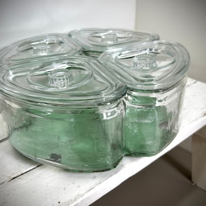 May include: Four clear glass storage jars with matching lids. The jars are arranged in a cloverleaf shape and sit on a white, distressed wooden surface. The jars have a slight green tint and are empty.
