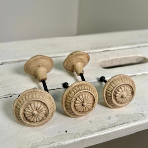 May include: Four antique-style cabinet knobs with a distressed, off-white finish. Each knob features a circular design with a central floral pattern and a black stem. The knobs are arranged on a white, weathered surface.