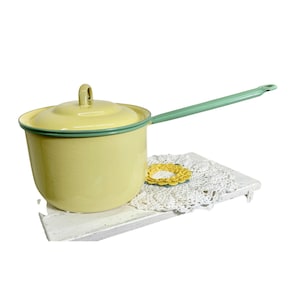 May include: A vintage yellow enamel saucepan with a lid and a long, mint green handle. The rim and handle are also mint green. The saucepan is sitting on a white, distressed wooden surface with a white lace doily and a yellow crocheted flower.