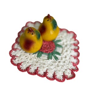 May include: A pair of vintage yellow bird-shaped salt and pepper shakers with red and green accents. They rest on a white crocheted doily with a red scalloped edge and a central red flower.