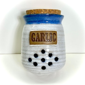 May include: A ceramic garlic keeper with a blue rim and a cork lid. The white body has a textured surface with black dot accents and a rectangular label that reads "GARLIC". A kitchen storage container.
