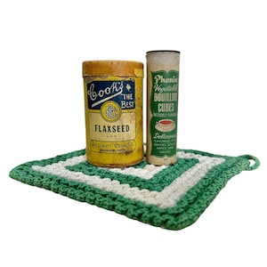 May include: A vintage still life featuring two antique food containers and a crocheted trivet. One container is a yellow can of "Books The Best Flaxseed" and the other is a cylindrical container of "Phenix Vegetable Bouillon Cubes". The trivet is green and white.