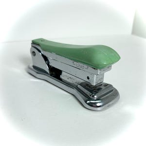 May include: A vintage stapler with a chrome body and a light green top. The word "LOAD" is printed on the side. The stapler is designed for office or school use.