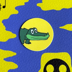 May include: A cartoon alligator illustration on a yellow circle, set against a blue and yellow patterned background. The alligator is green with a white belly and large, round eyes. The background has a musical note.