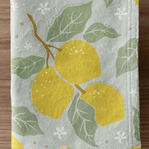 Peut inclure: Une serviette de toilette bleu clair avec un motif de branche de citron. La serviette présente des citrons jaunes, des feuilles vertes et de petites fleurs blanches. La serviette est suspendue à une barre argentée contre un mur en lambris.