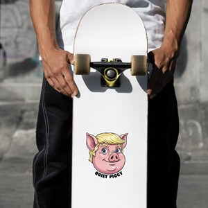 May include: White skateboard with a cartoon pig face sticker. The pig has blonde hair, blue eyes, and pink ears. The text "QUIET PIGGY" is below the pig's face. The skateboard is held by a person.
