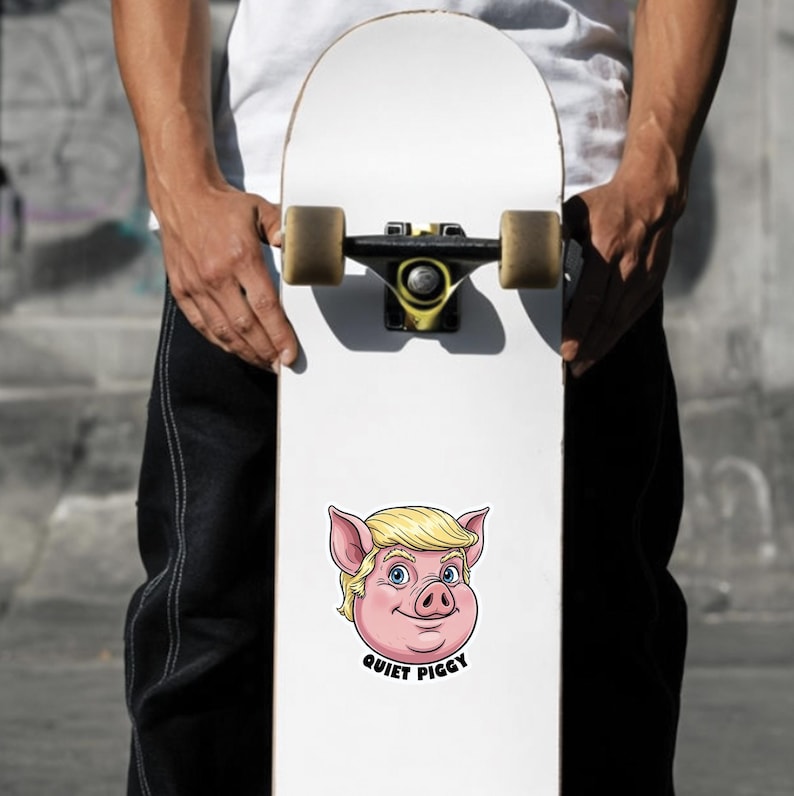 May include: White skateboard with a cartoon pig face sticker. The pig has blonde hair, blue eyes, and pink ears. The text "QUIET PIGGY" is below the pig's face. The skateboard is held by a person.