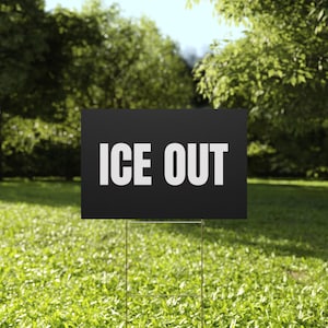 May include: A black sign with the words "ICE OUT" in bold white letters. The sign is placed in a grassy yard with trees in the background. The sign is held up by a metal wire frame.