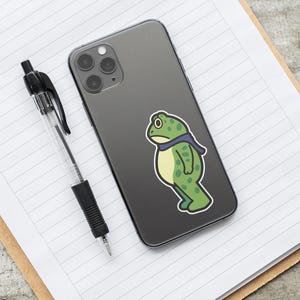 May include: A black smartphone with a green frog sticker and a black pen on a lined notepad. The frog sticker is green with a purple scarf. The pen is black and clear.