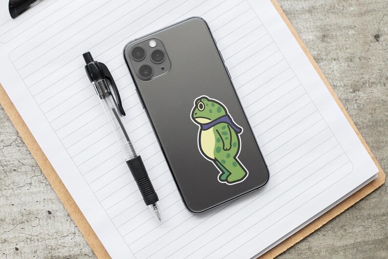 May include: A black smartphone with a green frog sticker and a black pen on a lined notepad. The frog sticker is green with a purple scarf. The pen is black and clear.
