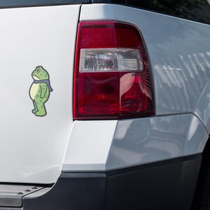 May include: A green cartoon frog sticker with a blue scarf is affixed to the white exterior of a vehicle. The frog has a sad expression. The vehicle's red taillight is visible.