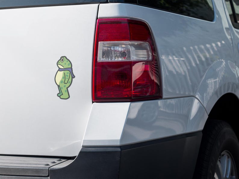 May include: A green cartoon frog sticker with a blue scarf is affixed to the white exterior of a vehicle. The frog has a sad expression. The vehicle's red taillight is visible.