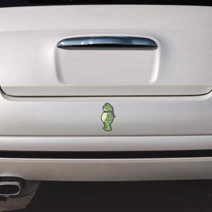 May include: A green frog sticker with a blue scarf is affixed to the rear of a white car. The sticker is centered on the car's trunk, below the chrome handle. The car has red taillights.