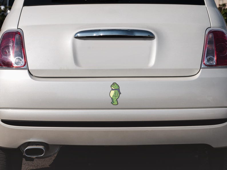 May include: A green frog sticker with a blue scarf is affixed to the rear of a white car. The sticker is centered on the car's trunk, below the chrome handle. The car has red taillights.
