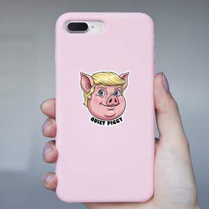May include: A pink phone case featuring a cartoon pig with blonde hair and the text "QUIET PIGGY" in black. The pig has pink skin, blue eyes, and a smiling expression. The phone case is held in a hand.