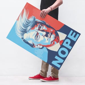 May include: A poster with a portrait in red, blue, and cream tones, with the word "NOPE" in blue. The poster is held by a person wearing a black top and beige trousers. The poster's design is bold and eye-catching.
