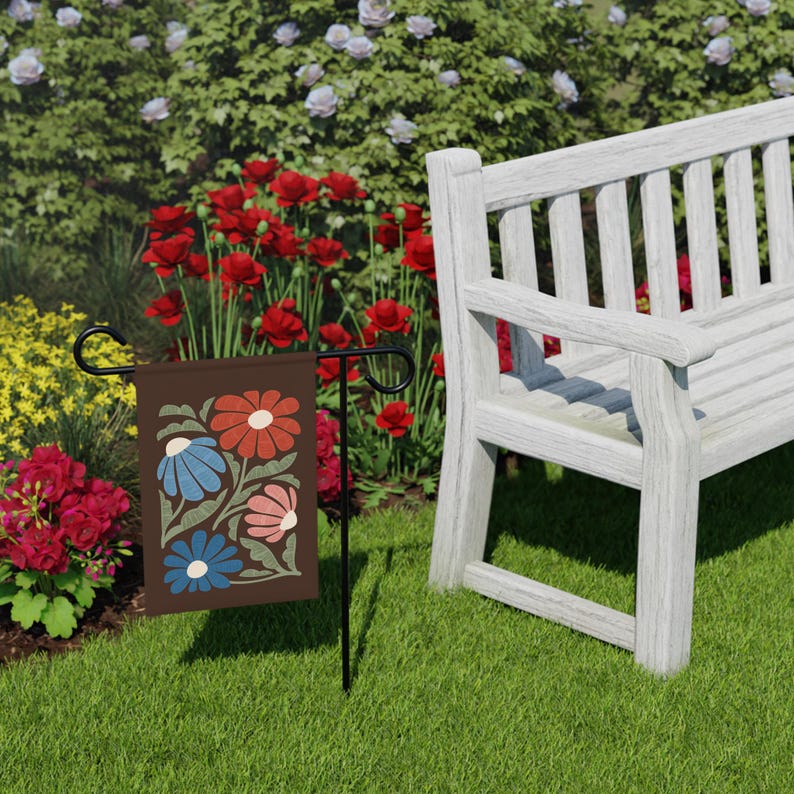 May include: A brown garden flag featuring a floral design in red, blue, and pink. The flag is displayed on a black metal stand in a garden setting with a white bench and colourful flowers.
