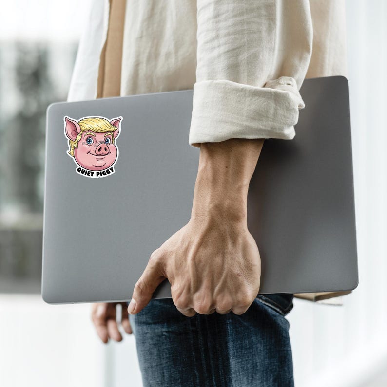 May include: A sticker featuring a cartoon pig with blonde hair and blue eyes. The pig has a pink face and is smiling. The text "QUIET PIGGY" is below the pig's face. The sticker is on a gray laptop.