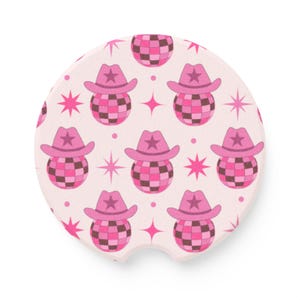 May include: A round car coaster with a repeating pattern of pink cowboy hats with star accents atop disco balls. The background is a light pink with pink starbursts. The coaster has a small notch for easy removal.