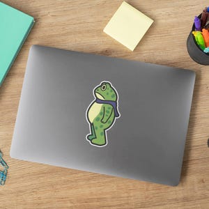 May include: A green frog sticker with a blue scarf is on a gray laptop. The frog has a sad expression. Other items on the wooden surface include a notepad, sticky note, pens, and a camera.