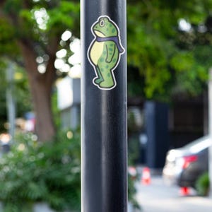 May include: A sticker of a cartoon frog with a purple scarf is affixed to a black pole. The frog is green with darker green spots and a yellow belly. The background is blurred, showing greenery and a street scene.