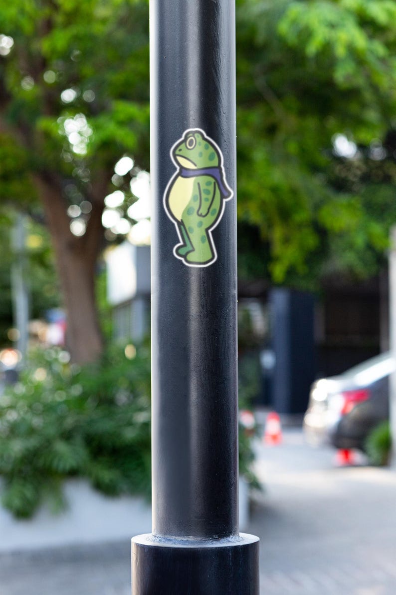 May include: A sticker of a cartoon frog with a purple scarf is affixed to a black pole. The frog is green with darker green spots and a yellow belly. The background is blurred, showing greenery and a street scene.
