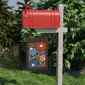 May include: A red mailbox with its flag raised, fixed to a wooden post. A brown garden flag hangs beneath, displaying a floral design with red, blue, and pink flowers. The flag is set in a garden with green grass and foliage.