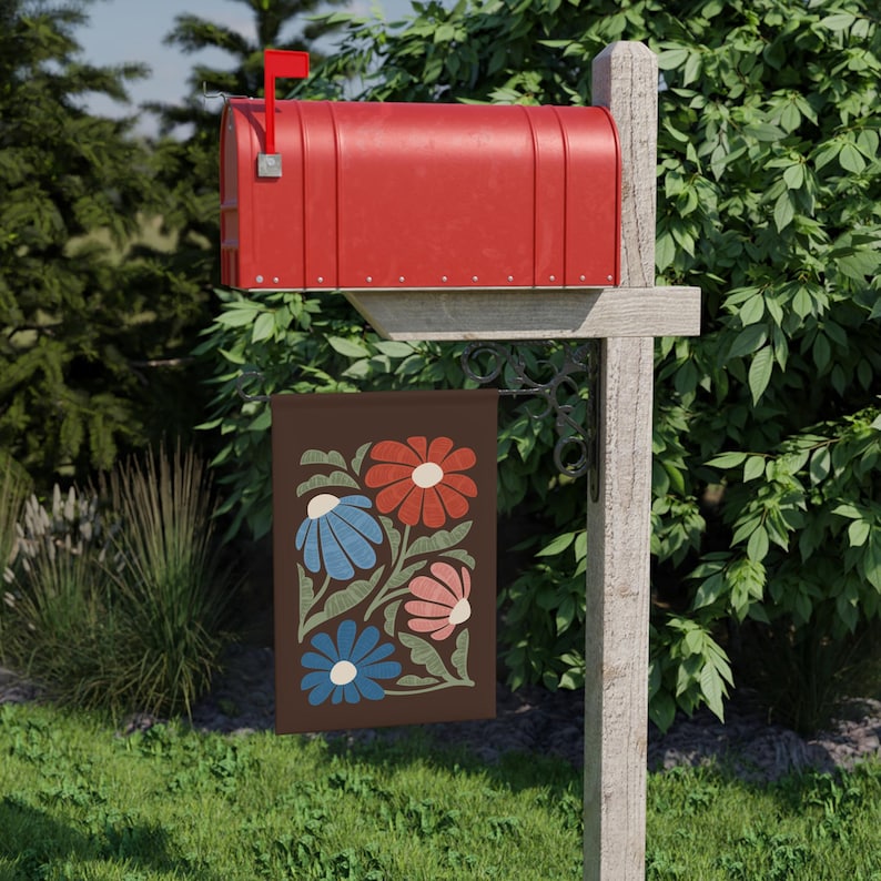 May include: A red mailbox with its flag raised, fixed to a wooden post. A brown garden flag hangs beneath, displaying a floral design with red, blue, and pink flowers. The flag is set in a garden with green grass and foliage.