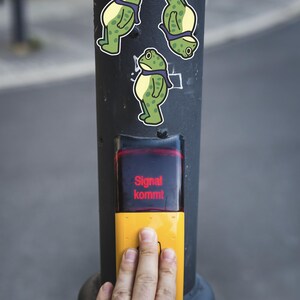 May include: A black pole with three frog stickers and a yellow pedestrian crossing button. The button has the text "Signal kommt" in red. A hand is pressing the button.