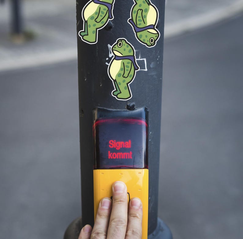 May include: A black pole with three frog stickers and a yellow pedestrian crossing button. The button has the text "Signal kommt" in red. A hand is pressing the button.