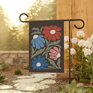 May include: A dark brown garden flag featuring a floral design with red, blue, and pink flowers. The flowers have white centers and are set against green leaves. The flag is displayed on a black metal stand in a garden setting.