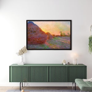 May include: Framed artwork showing a sunset over haystacks in a field. The painting uses warm colours of orange, yellow, and red, with haystacks in the foreground and trees in the distance. The artwork is displayed above a green cabinet.