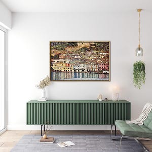 Op de afbeelding: Een schilderij van een kleurrijk stadsbeeld met gebouwen langs een waterkant. De gebouwen zijn geschilderd in tinten geel, oranje, roze en groen. Het water is diepblauw en reflecteert de gebouwen. Het schilderij is ingelijst in een houten lijst.