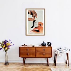 May include: A framed surrealist art print featuring a large hand holding a tiny person above a giant open mouth. The artwork uses a collage style with realistic skin tones, a black silhouette figure, and a light background, all encased in a wooden frame.